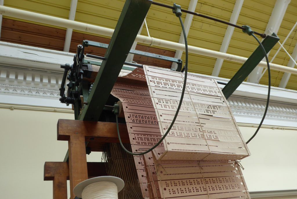 The punch cards used by the Jacquard loom are pictured. Long strips of card are sewn together, each card has a sequence of holes in it. The holes in the card correspond to a line of pattern in the woven fabric.